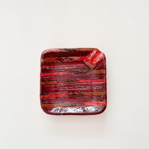 May include: Square ceramic ashtray with a red, brown, and black striped pattern. The ashtray has a small, raised corner for resting cigarettes. The design features horizontal stripes in varying shades of red and brown.