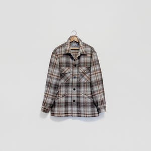 May include: A plaid button-up shirt in shades of brown, gray, and beige hangs on a wooden hanger. The shirt features two chest pockets and a classic collar. The fabric has a textured appearance.