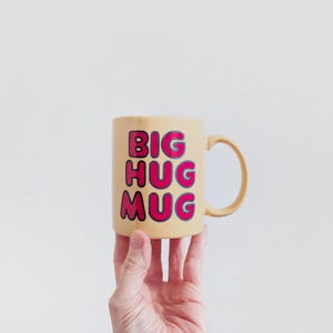 May include: A beige ceramic mug with the words "BIG HUG MUG" in pink block letters. The mug is held up by a hand against a white background. The mug has a curved handle.