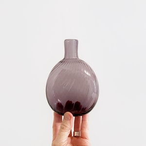 May include: A small, round, purple glass vase with a narrow neck. The vase has a textured, ribbed design on the body and a dark purple base. The vase is held by a hand against a white background.