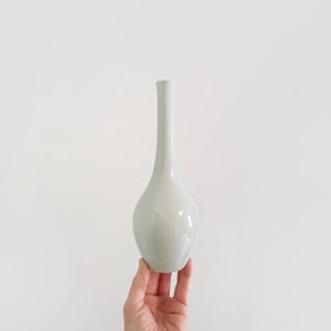 以下が含まれることがあります： 細身の首と丸みを帯びたボディを持つ白い陶器の花瓶。花瓶はシンプルでエレガントなデザインです。