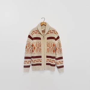 May include: A cream-colored zip-up cardigan sweater with a shawl collar. The sweater features geometric patterns in shades of burgundy and orange. The sweater is hanging on a wooden hanger.