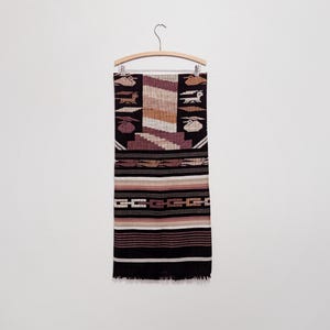 May include: A rectangular woven textile hanging on a wooden hanger. The fabric features a black background with horizontal bands of tan, brown, and cream. It displays repeating patterns of stylized animals and geometric shapes in muted earth tones.