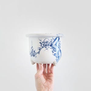 May include: White ceramic planter with blue floral and bird design. The planter has three small feet and a wide rim. The blue design is hand-painted and covers the exterior of the planter. A decorative piece for plants.