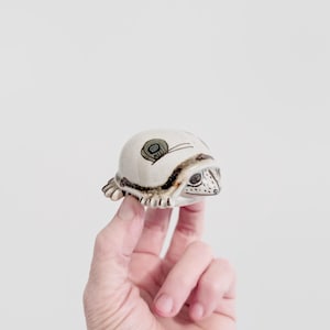 May include: A cream-coloured ceramic turtle figurine, featuring a snail design on its shell. The turtle has black and brown detailing, a detailed face, and small feet. The figurine is held up by a hand against a white background.