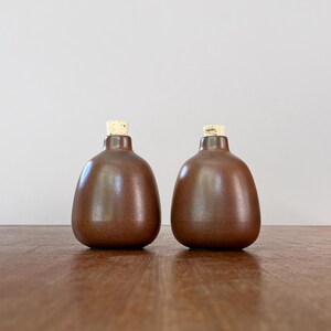 May include: Two brown ceramic bottles with cork stoppers. The bottles have a rounded shape and a smooth, glossy finish. The corks are light brown. The bottles are displayed on a wooden surface.