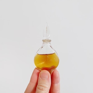May include: A small, clear glass perfume bottle with a teardrop-shaped body and a long, pointed stopper. The bottle is filled with a golden-colored liquid. The word "JUMADE" is printed on the stopper. The bottle is held by a person.