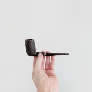Peut inclure: Une pipe à tabac marron foncé avec un foyer texturé et une tige lisse et fine. La pipe a une bande dorée près de la jonction de la tige et du foyer. La pipe est tenue sur un fond blanc uni.