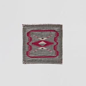 May include: A small, square gray textile with a geometric design. The design features a central diamond shape in white and pink, framed by a pink border and repeating patterns. The edges are frayed.