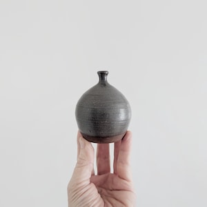 May include: A small, round ceramic bud vase with a narrow neck and a dark, metallic-grey glazed finish. The base of the vase is unglazed, revealing a natural, earthy brown clay texture that contrasts with the smooth, reflective upper body.