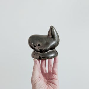 May include: A small, smooth, dark gray ceramic sculpture with a rounded, abstract form. The object features a glossy, reflective surface that catches the light, creating highlights across its curved, organic shape. It sits on a circular base.