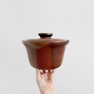 May include: A brown ceramic casserole dish with a lid. The dish has a rounded shape and a dark brown handle on the lid. The exterior has a glossy finish with a gradient of brown hues.