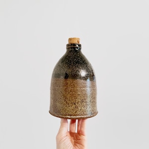 May include: A handcrafted ceramic bottle with a cork stopper. The bottle has a speckled brown and black glaze. The rounded body transitions to a narrower neck, topped with a natural cork. The bottle is a rustic, decorative item.