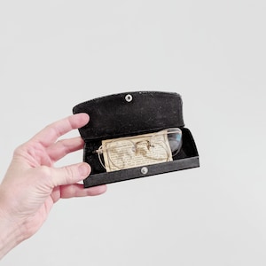 May include: A black eyeglass case with a pair of vintage eyeglasses inside. The glasses have thin, gold-colored frames and clear lenses. A small, rectangular paper with text is also inside the case.