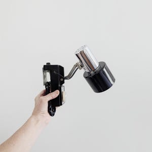 May include: A wall-mounted lamp featuring a black rectangular base with a circular control knob and a black power cord. The lamp head is a cylindrical chrome and black metal fixture attached by an adjustable metal arm for directional lighting.