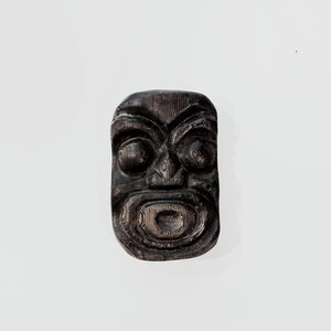 May include: A dark brown, rectangular wooden mask featuring deeply carved facial features, including prominent rounded eyes, a defined nose, and an open, oval-shaped mouth. The surface displays a visible wood grain texture throughout the carving.