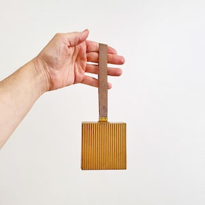 May include: A vintage gold-toned rectangular clutch with vertical ribbed detailing. It features a matching brown strap and a small gold-colored clasp. The clutch is held up by a hand against a white background.