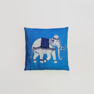 Pode incluir: Uma almofada quadrada de cor azul vibrante com um design central bordado de um elefante branco. O elefante est&aacute; adornado com uma manta azul-escura estampada, um adorno de cabe&ccedil;a decorativo e detalhes intrincados nas patas e na tromba.