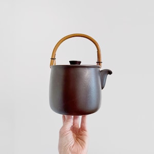 May include: A dark brown ceramic teapot with a smooth, matte finish, featuring a rounded body, a small lid, a short spout, and a curved, light-colored wooden handle attached to two metal loops on the top rim of the vessel.