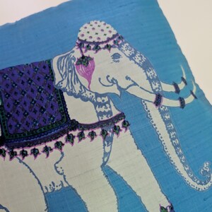 Pode incluir: Uma capa de almofada quadrada de tecido azul com uma ilustra&ccedil;&atilde;o de um elefante branco. O elefante est&aacute; adornado com uma manta roxa estampada, um adorno de cabe&ccedil;a decorativo e faixas nas patas. O design inclui detalhes em roxo e verde.