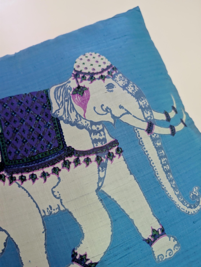 Pode incluir: Uma capa de almofada quadrada de tecido azul com uma ilustra&ccedil;&atilde;o de um elefante branco. O elefante est&aacute; adornado com uma manta roxa estampada, um adorno de cabe&ccedil;a decorativo e faixas nas patas. O design inclui detalhes em roxo e verde.