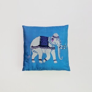 Pode incluir: Uma almofada quadrada de cor azul vibrante com um gr&aacute;fico central de um elefante branco. O elefante est&aacute; adornado com uma manta estampada em azul escuro, um adorno de cabe&ccedil;a decorativo e detalhes em azul escuro nas pernas e no corpo.
