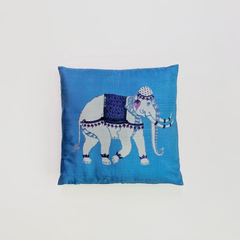 Pode incluir: Uma almofada quadrada de cor azul vibrante com um gr&aacute;fico central de um elefante branco. O elefante est&aacute; adornado com uma manta estampada em azul escuro, um adorno de cabe&ccedil;a decorativo e detalhes em azul escuro nas pernas e no corpo.