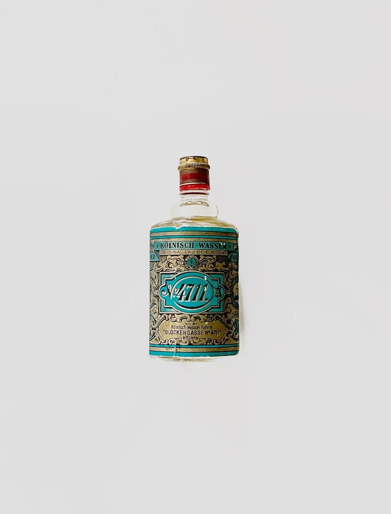 Puede incluir: Una botella de colonia de vidrio transparente con una etiqueta dorada y azul. La etiqueta dice "K&Ouml;LNISCH WASSER FABRIK "GLOCKENGASSE N&deg;4711" K&Ouml;LN".