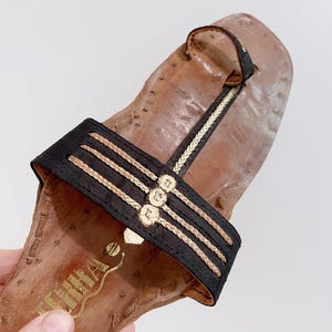 以下が含まれることがあります： 黒いストラップと金色のアクセントが付いた茶色の革製サンダル。サンダルはスクエアトゥとトングストラップが特徴です。黒いストラップには、金色の編み込みのディテールと装飾的な金の装飾が施されています。サンダルは手で持たれています。
