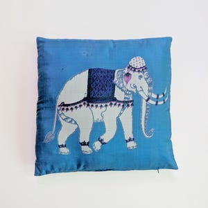 Pode incluir: Uma almofada quadrada de cor azul vibrante com um gr&aacute;fico central de um elefante branco. O elefante est&aacute; adornado com uma manta azul-escura estampada, um adorno de cabe&ccedil;a decorativo e detalhes em roxo nas orelhas e nas presas.