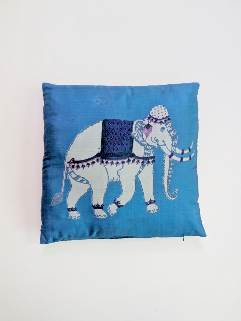 Pode incluir: Uma almofada quadrada de cor azul vibrante com um gr&aacute;fico central de um elefante branco. O elefante est&aacute; adornado com uma manta azul-escura estampada, um adorno de cabe&ccedil;a decorativo e detalhes em roxo nas orelhas e nas presas.