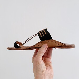 以下が含まれることがあります： 茶色のソールと黒のアクセントが特徴の手作りの革製サンダル。 サンダルは、つま先ループと足の甲にストラップが付いています。 デザインには装飾的なステッチとミニマリストな美学が含まれています。