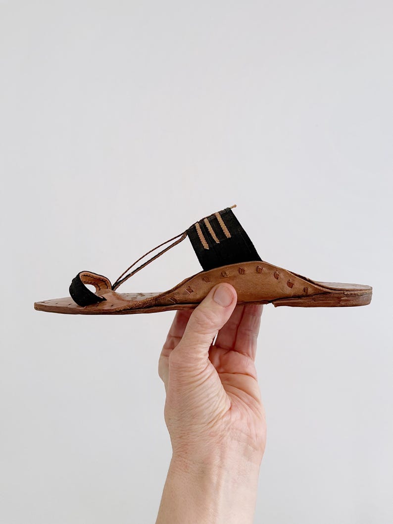 以下が含まれることがあります： 茶色のソールと黒のアクセントが特徴の手作りの革製サンダル。 サンダルは、つま先ループと足の甲にストラップが付いています。 デザインには装飾的なステッチとミニマリストな美学が含まれています。