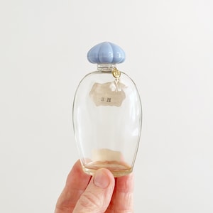 May include: A clear glass perfume bottle with a light blue, pumpkin-shaped stopper. The bottle has a paper label with the text "3 H" and a gold-colored charm. The bottle is held by a hand.