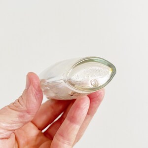 May include: A clear, oval-shaped glass bottle with a rounded base and a slightly pointed top. The bottle is held in a hand, and a small label is visible near the base. The bottle has a small, circular marking on the bottom.