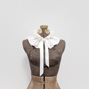 May include: A vintage-style white ruffled collar with a long white ribbon, displayed on a brown dress form. The dress form has a tan-colored skirt. The background is plain white.