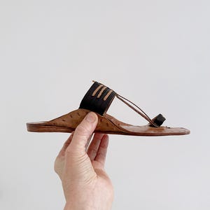 以下が含まれることがあります： 茶色の革製サンダル、トングストラップのデザイン。サンダルは、上部に濃い茶色のストラップとつま先ループが付いています。ソールは明るい茶色です。サンダルは手で持たれています。