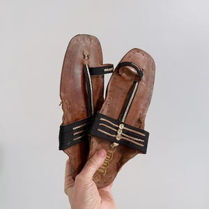 以下が含まれることがあります： 黒いストラップと金色のアクセントが付いた、茶色の革製サンダルのペア。 サンダルはトングスタイルのデザインで、手で持っています。 サンダルにはサイズ7のラベルが付いています。