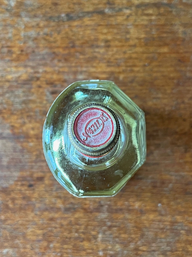 Puede incluir: Una botella de vidrio transparente con una tapa roja y dorada. La tapa tiene la palabra "JAML" impresa.