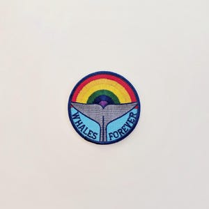 May include: Embroidered patch with a rainbow and whale tail design. The patch features a blue border with the words "Whales Forever" in white. The whale tail is gray, and the rainbow has multiple colors.