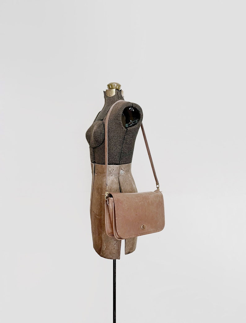 May include: A brown leather shoulder bag with a gold clasp and a long strap. The bag is hanging on a dress form.
