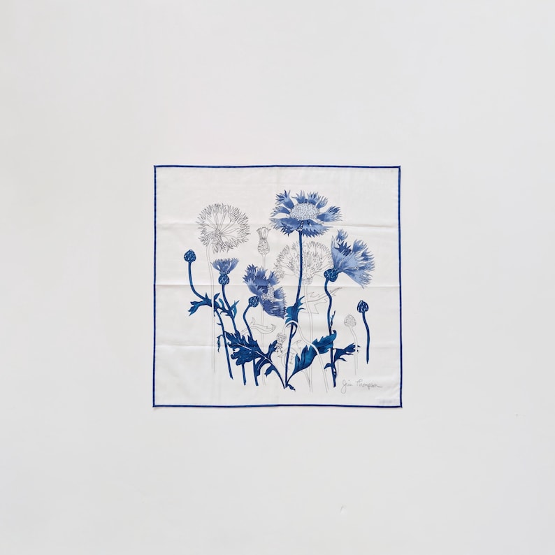 Puede incluir: Un pa&ntilde;uelo de seda blanco cuadrado con un estampado bot&aacute;nico azul de varias flores silvestres y cardos. La tela tiene un borde azul fino en los cuatro lados e incluye una firma en la esquina inferior derecha de la ilustraci&oacute;n.
