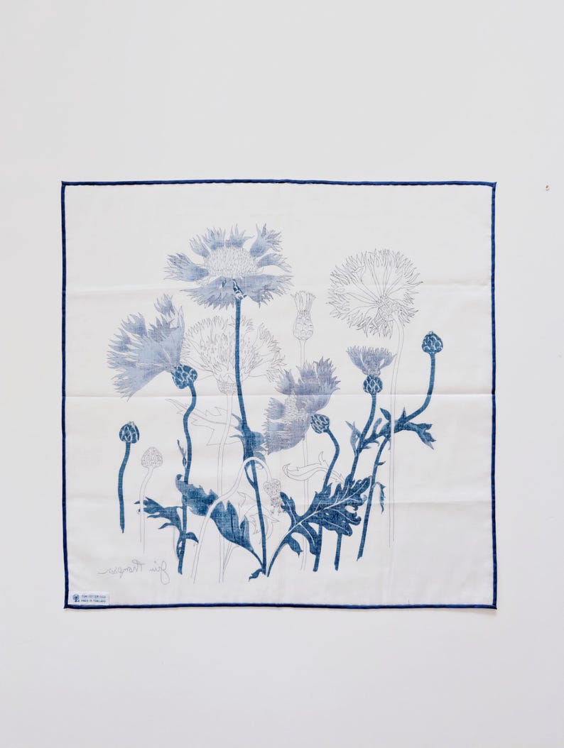 Puede incluir: Un pa&ntilde;uelo de bolsillo cuadrado de color blanco con un estampado bot&aacute;nico azul de acianos y cardos. La tela tiene un borde fino de color azul en los cuatro lados. Una peque&ntilde;a etiqueta blanca es visible en la esquina inferior izquierda.