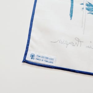 Puede incluir: Un textil de algod&oacute;n blanco con un borde azul y una ilustraci&oacute;n bot&aacute;nica azul. Una etiqueta de tela blanca est&aacute; cosida en la esquina, impresa con un icono de rueca y el texto: THAI COTTON 100% MADE IN THAILAND.