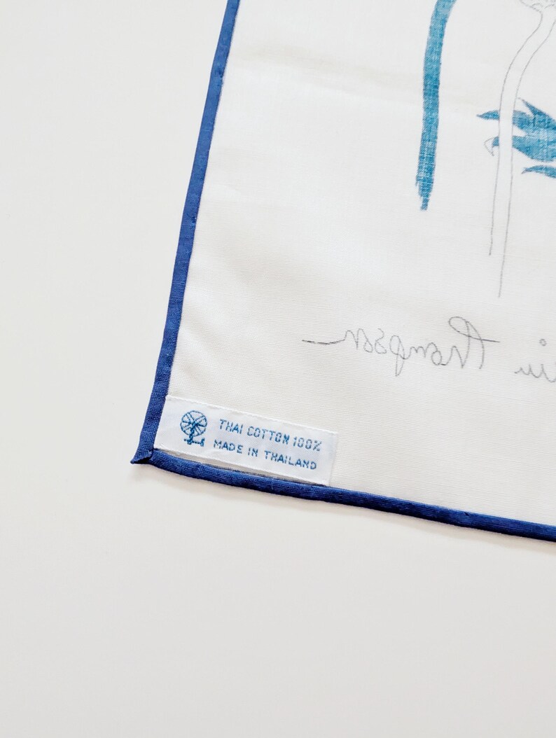 Puede incluir: Un textil de algod&oacute;n blanco con un borde azul y una ilustraci&oacute;n bot&aacute;nica azul. Una etiqueta de tela blanca est&aacute; cosida en la esquina, impresa con un icono de rueca y el texto: THAI COTTON 100% MADE IN THAILAND.