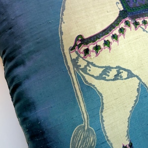 Pode incluir: Uma capa de almofada de seda azul com uma ilustra&ccedil;&atilde;o impressa de um elefante na cor creme. O elefante &eacute; adornado com uma manta estampada em roxo e verde nas costas, juntamente com um acabamento decorativo rosa e verde na lateral e nas patas.
