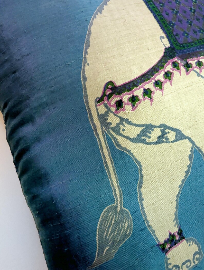 Pode incluir: Uma capa de almofada de seda azul com uma ilustra&ccedil;&atilde;o impressa de um elefante na cor creme. O elefante &eacute; adornado com uma manta estampada em roxo e verde nas costas, juntamente com um acabamento decorativo rosa e verde na lateral e nas patas.