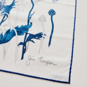 Puede incluir: Un pa&ntilde;uelo cuadrado blanco con un estampado floral azul de acianos y tallos. El dise&ntilde;o incluye ilustraciones bot&aacute;nicas detalladas en azul y contornos. El nombre Jim Thompson est&aacute; impreso en letra negra. El pa&ntilde;uelo tiene un dobladillo azul.