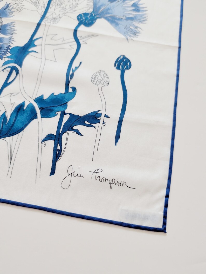 Puede incluir: Un pa&ntilde;uelo cuadrado blanco con un estampado floral azul de acianos y tallos. El dise&ntilde;o incluye ilustraciones bot&aacute;nicas detalladas en azul y contornos. El nombre Jim Thompson est&aacute; impreso en letra negra. El pa&ntilde;uelo tiene un dobladillo azul.
