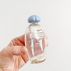 May include: A clear glass perfume bottle with a light blue cap. The bottle has a label with the words "Quelques Fleurs Houbigant" and "Eau de Toilette". The bottle is held in a hand.